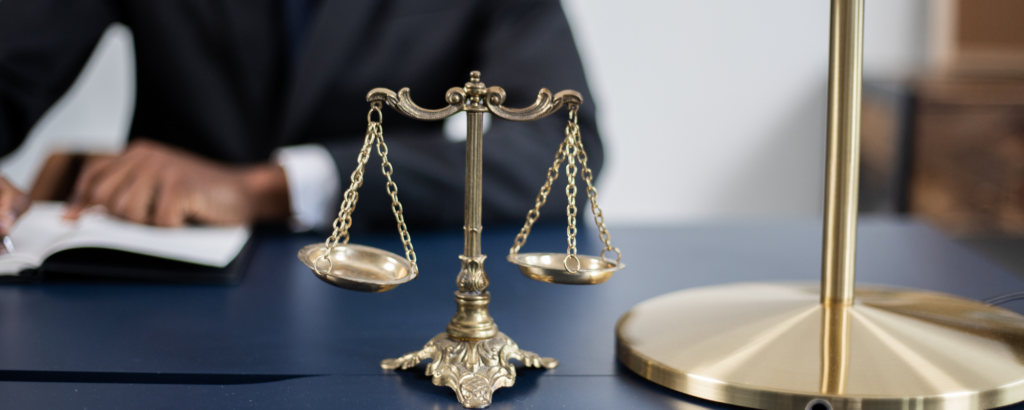 Image of a lawyer sitting at a desk, writing on legal documents. Gold scales of Justice are nearby.
