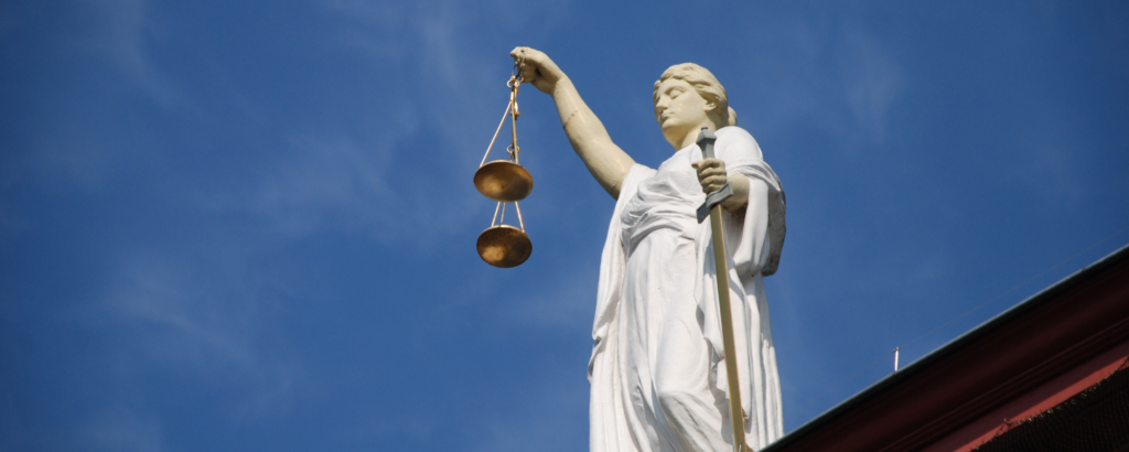 A large white statue of Lady Justice, outside with a blue sky as the background.