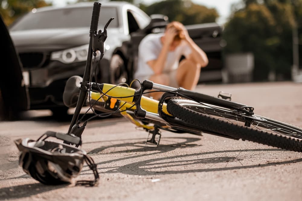 A fallen yellow bicycle in the foreground with a blurred person seated on the ground in the background, near a car.
