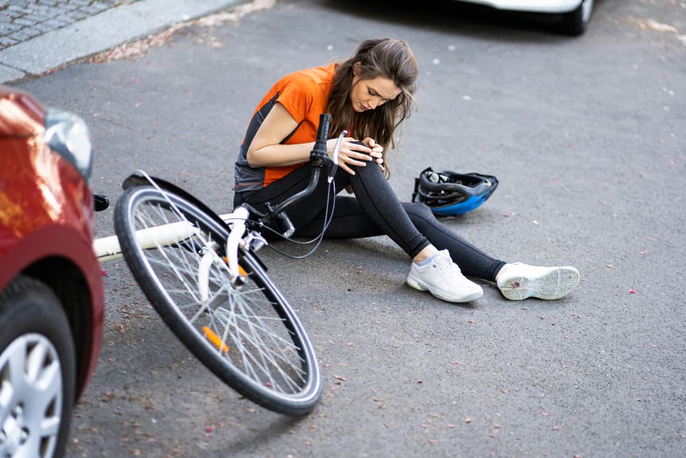 Injured cyclist sitting on the ground checking her knee near an overturned bicycle and a red car, with helmet aside.