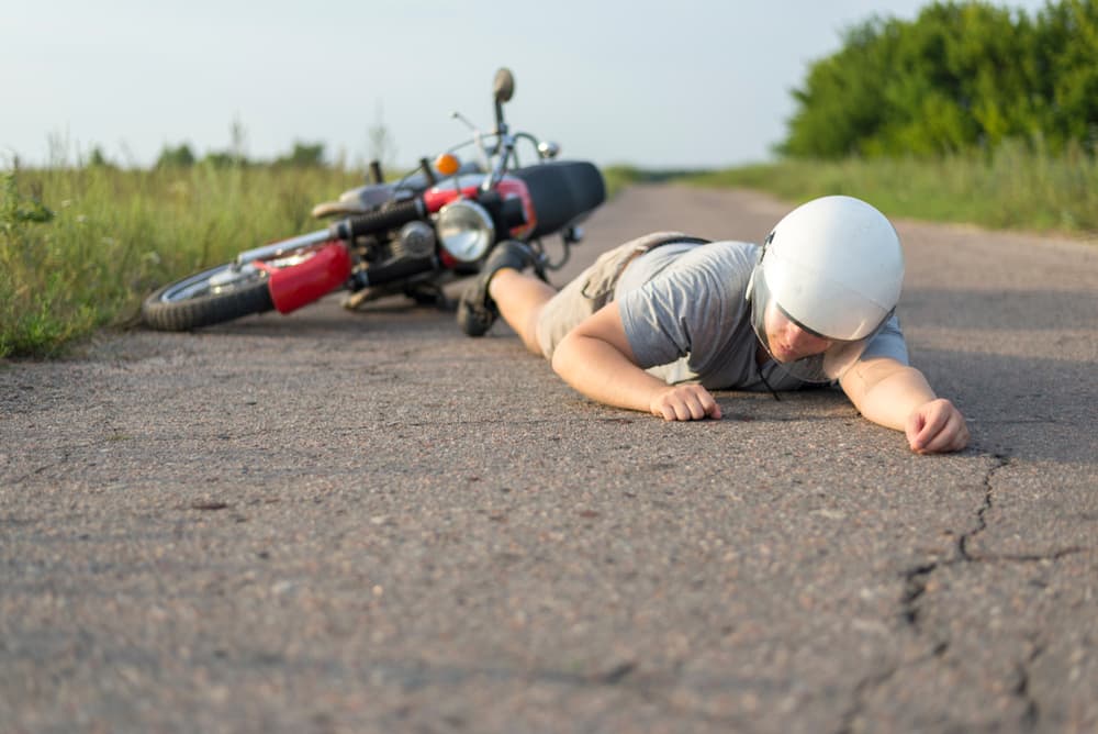 Motorcyclist lying on the ground after an accident with their motorcycle tipped over in the background on a rural road.