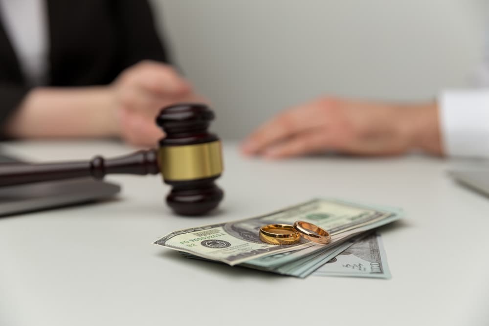 A wooden gavel on a desk next to two gold wedding rings on a stack of dollar bills, implying a divorce settlement.