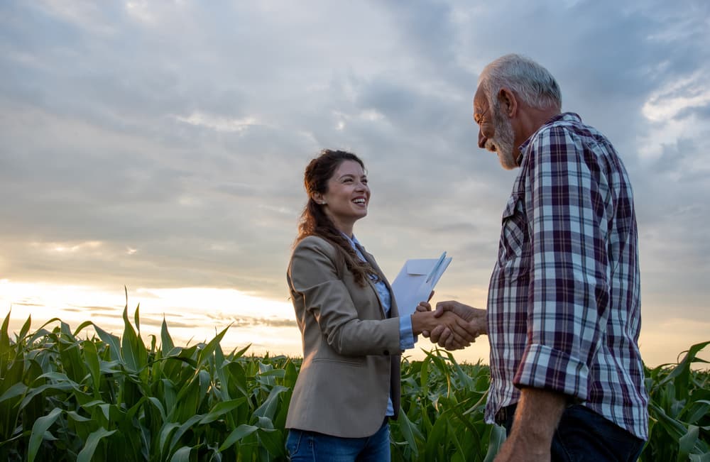 Senior farmer shaking hands with a young woman holding a notebook in a cornfield. Agribusiness insurance concept.