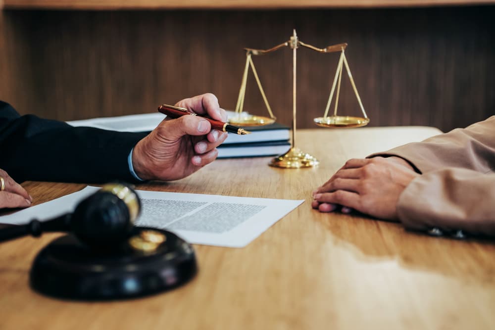 Two individuals in a legal setting with a pen, document, gavel, and scales of justice on a wooden table.