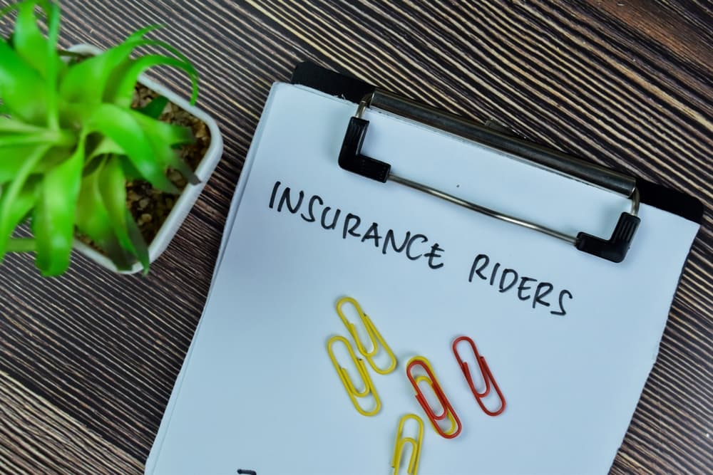 Clipboard with the words "INSURANCE RIDERS" written on it, paperclips, and a green plant on a wooden background.