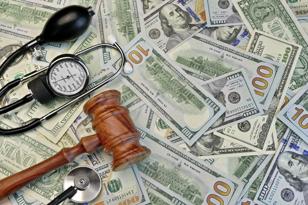 A wooden gavel, stethoscope, and blood pressure monitor lay atop a scattering of various US dollar bills.