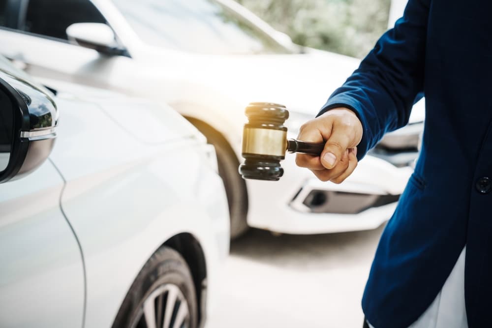 A lawyer holding a wooden gavel stands in front of a car, discussing legal issues such as citations, liability, and negligence related to traffic violations.