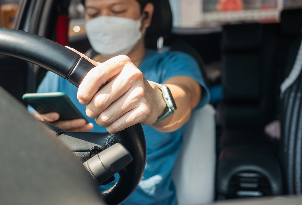 Close-up of a masked driver looking at a smartphone while holding the steering wheel, illustrating distracted driving.