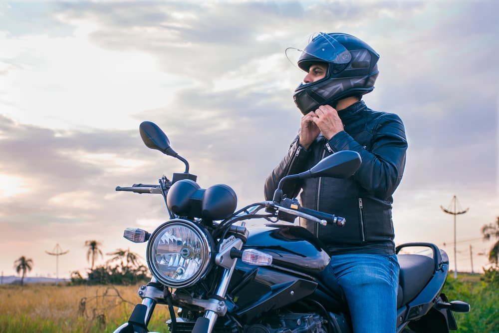 Man sitting on a motorcycle, wearing jeans and a black jacket, fastening his helmet with a landscape in the background.