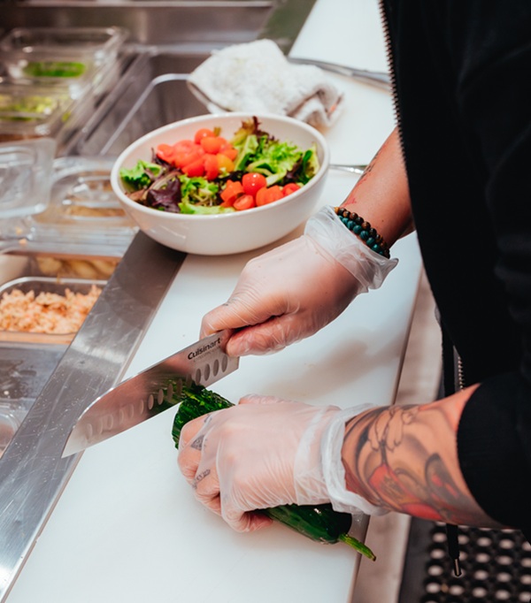 A person making salad, while cutting cucumber at a busy food court table