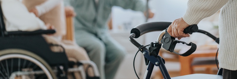 Closeup of an elderly woman using a walker, with other residents blurred in the background.